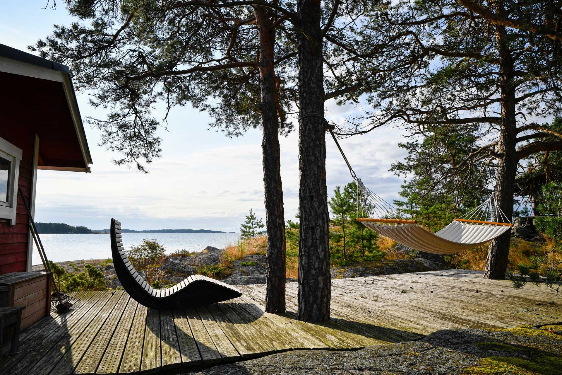 Äldre terrass vid en röd stuga. Tallar, klippor, hav.